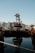 A red and black industrial structure bearing the name 'SUMITOMO' is situated on water. Surrounding it are various other industrial buildings and equipment. The calm water reflects the scene, and tall buildings with mountainous terrain in the background can be seen under a clear blue sky.
