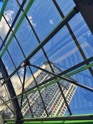A modern architectural structure displays a reflection of a high-rise building on its glass surface. The angular lines of the metal framework create a geometric pattern, and the glass has a blue tint, revealing a partial view of clouds and the sky.