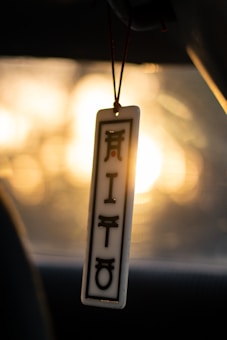 A rectangular pendant with stylized, black symbols hangs from a thread against the backdrop of a softly glowing sunset. The pendant features a small red dot among the symbols and is framed by a dark border. The background is blurred, hinting at the interior of a vehicle with the light creating a serene bokeh effect.