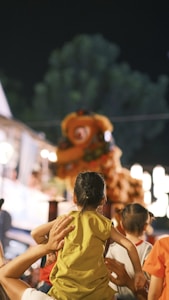A child wearing a yellow shirt is being held up, facing a colorful lion dance performance during a festival at night. The child is surrounded by other spectators, with bright lights and blurred figures in the background.