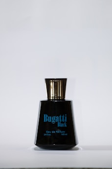 A black perfume bottle with a shiny gold cap. The label reads 'Bugatti Black' along with details about the volume and type of product in blue text.