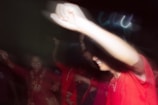 A group of people appears to be dancing with joyful expressions. The image is slightly blurred, suggesting movement or excitement. The main focus is on an individual wearing a bright red shirt, with their arm raised in celebration. Other individuals in similar bright attire are visible in the background, enhancing the festive atmosphere.