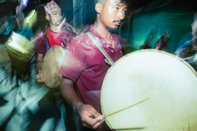 A group of men, dressed in casual clothing, are playing large traditional drums. The man in the foreground is focusing on his performance, while the others behind him also concentrate on their drumming. The scene seems to be in motion, as indicated by the blur effect around the figures and drums.