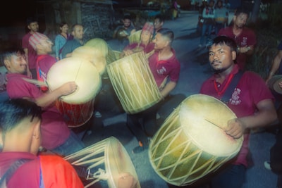 A group of people engages in a lively drum performance outdoors, with most participants wearing maroon shirts. The drummers carry large traditional drums, and their expressions are focused and enthusiastic. The scene is dimly lit, suggesting an evening or nighttime event. Spectators and other performers stand in the background, adding to the festive atmosphere.