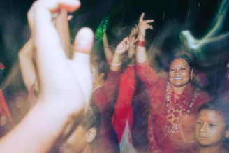A group of people, mostly women, are dancing and celebrating. They are wearing traditional clothing and have joyful expressions on their faces. There are several raised hands and colorful outfits, indicating a lively and festive atmosphere.