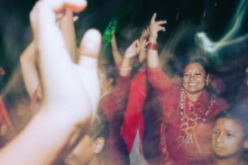 A group of people, mostly women, are dancing and celebrating. They are wearing traditional clothing and have joyful expressions on their faces. There are several raised hands and colorful outfits, indicating a lively and festive atmosphere.