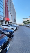 A line of parked cars along a street beside a tall commercial building. The building has multiple signs and logos, suggesting various businesses, including fitness centers and restaurants. The sky is clear and blue, indicating a sunny day.