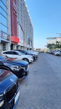 A line of parked cars along a street beside a tall commercial building. The building has multiple signs and logos, suggesting various businesses, including fitness centers and restaurants. The sky is clear and blue, indicating a sunny day.
