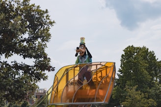 A tall character dressed in bright colors stands on a large, yellow-tinted transparent platform. Surrounding the character are lush green trees against a partly cloudy sky.