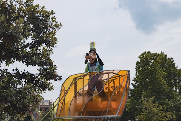 A tall character dressed in bright colors stands on a large, yellow-tinted transparent platform. Surrounding the character are lush green trees against a partly cloudy sky.