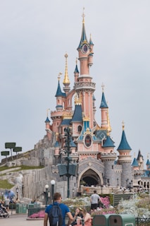A whimsical castle with pink walls and blue spires adorned with gold accents stands prominently. The architecture is fairytale-like with intricate details and lush greenery surrounding it. Visitors are seen around the castle, enjoying the vibrant and enchanting atmosphere.