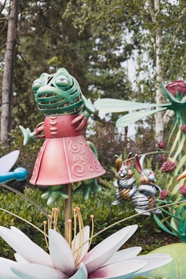 A whimsical sculpture garden featuring a green, anthropomorphic frog dressed in a pink, ornate outfit, standing on a large white flower. In the background, there are two stylized, cartoonish rabbit sculptures and other oversized, colorful botanical elements. Tall trees and lush greenery surround the area.
