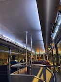 The interior of a dimly lit bus with several passengers seated. The lighting creates a warm and intimate ambiance, with reflections visible on the bus windows. There are metal poles for standing passengers and red cushioned seats arranged in rows.