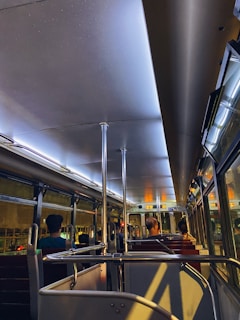 The interior of a dimly lit bus with several passengers seated. The lighting creates a warm and intimate ambiance, with reflections visible on the bus windows. There are metal poles for standing passengers and red cushioned seats arranged in rows.