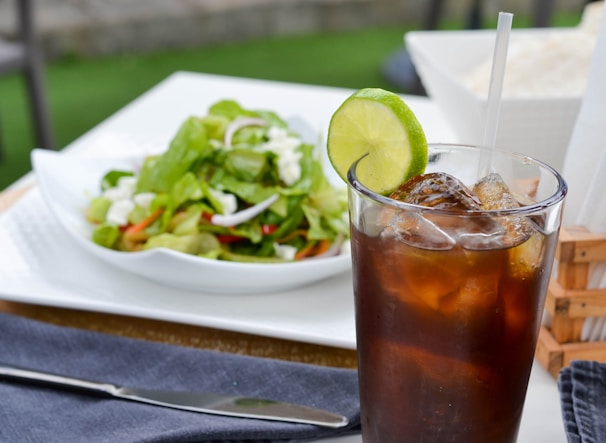 A chilled glass of cola with a slice of lime, set against a backdrop of colorful Pakistani textiles.