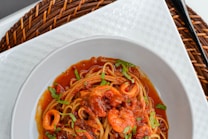 A bowl of spaghetti is topped with a rich tomato-based seafood sauce, featuring shrimp and garnished with green herbs. The dish is presented on a white plate atop a woven textured charger.