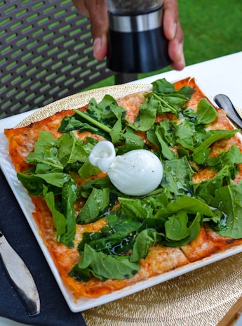 A square-shaped pizza topped with fresh arugula and a soft, round burrata cheese placed at the center. The pizza is placed on a white plate with a golden woven placemat underneath. A hand is seen using a pepper grinder above the pizza, ready to add seasoning. A partially visible knife and fork are placed beside the plate on a dark napkin, and a metal chair is visible in the background.