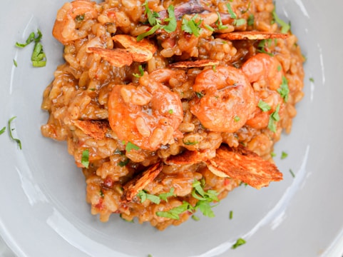 A plate of shrimp risotto garnished with fresh herbs and crispy chips. The dish has a creamy texture with visible pieces of shrimp and seasonings.