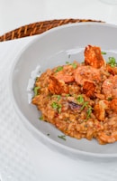 A gourmet dish featuring creamy rice mixed with shrimp and garnished with fresh herbs and crispy elements, served in an elegant grey bowl. The table setting includes a woven placemat beneath the bowl.
