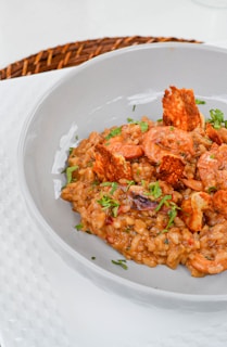 A gourmet dish featuring creamy rice mixed with shrimp and garnished with fresh herbs and crispy elements, served in an elegant grey bowl. The table setting includes a woven placemat beneath the bowl.