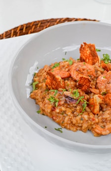 A gourmet dish featuring creamy rice mixed with shrimp and garnished with fresh herbs and crispy elements, served in an elegant grey bowl. The table setting includes a woven placemat beneath the bowl.