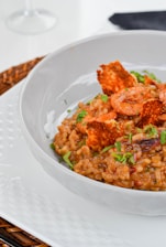 A white bowl contains a serving of risotto with shrimp and garnished with fresh herbs. The dish includes crispy toppings and appears creamy and well-cooked. The presentation is elegant, set on a white plate with a woven placemat underneath.