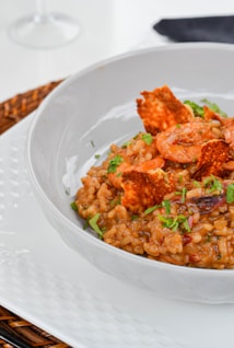 A white bowl contains a serving of risotto with shrimp and garnished with fresh herbs. The dish includes crispy toppings and appears creamy and well-cooked. The presentation is elegant, set on a white plate with a woven placemat underneath.