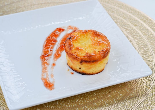 A small, baked dessert with a golden-brown crust is placed on a white, square plate. Next to the dessert, there is a drizzle of reddish-orange sauce, artfully arranged. The plate rests on a woven placemat, adding a textured background.