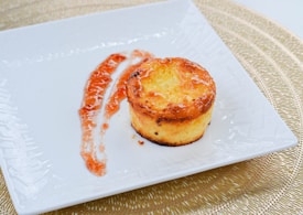 A small, baked dessert with a golden-brown crust is placed on a white, square plate. Next to the dessert, there is a drizzle of reddish-orange sauce, artfully arranged. The plate rests on a woven placemat, adding a textured background.