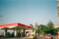 A gas station with a red and white canopy featuring the Caltex logo. Several cars, including a yellow taxi, are seen in the vicinity. There is a prominent signpost displaying fuel prices. Trees and a clear blue sky can be seen in the background.