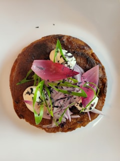 A beautifully plated dish featuring a base of a round, brown, crispy item topped with dollops of white cream. It is garnished with pink pickled onions, green herbs, and thin slices of red onion. There are also small flecks of a dark ingredient sprinkled on top.
