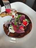 A gourmet dish presented on a white plate features a savory pancake topped with a dollop of white cream. Garnished with thinly sliced red onions, green herbs, and vibrant red flower petals, a fork holds a piece of the pancake with cream at the edge of the plate.