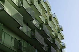 A series of modern architectural balconies with an overhanging design and uniform green color, arranged vertically on the facade of a building. The balconies feature metal mesh railings and are aligned in an alternating pattern, casting shadows and creating geometric shapes.