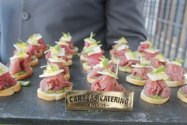 A tray of elegantly presented hors d'oeuvres with thin slices of raw meat, possibly beef, placed on small rounds of bread. Each piece is garnished with small greens, cheese slices, and a dollop of sauce. The arrangement is on a dark serving platter, and a sign with 'Caracas Catering Eventos' is prominently displayed in front.