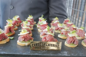 A tray of elegantly presented hors d'oeuvres with thin slices of raw meat, possibly beef, placed on small rounds of bread. Each piece is garnished with small greens, cheese slices, and a dollop of sauce. The arrangement is on a dark serving platter, and a sign with 'Caracas Catering Eventos' is prominently displayed in front.