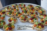 A platter of intricately arranged small tartlets, each topped with a colorful array of fresh fruits and vegetables, including grape tomatoes, green and orange melon balls, and garnished with greens. They are set on a round white tray with a label from a catering company.