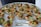 A platter of intricately arranged small tartlets, each topped with a colorful array of fresh fruits and vegetables, including grape tomatoes, green and orange melon balls, and garnished with greens. They are set on a round white tray with a label from a catering company.