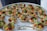 A platter of intricately arranged small tartlets, each topped with a colorful array of fresh fruits and vegetables, including grape tomatoes, green and orange melon balls, and garnished with greens. They are set on a round white tray with a label from a catering company.