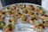 A platter of intricately arranged small tartlets, each topped with a colorful array of fresh fruits and vegetables, including grape tomatoes, green and orange melon balls, and garnished with greens. They are set on a round white tray with a label from a catering company.