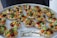 A platter of intricately arranged small tartlets, each topped with a colorful array of fresh fruits and vegetables, including grape tomatoes, green and orange melon balls, and garnished with greens. They are set on a round white tray with a label from a catering company.