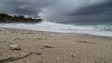 A stormy beach scene with dark clouds looming over the ocean. The waves crash onto a sandy shore, where small pebbles and debris are scattered. The atmosphere is tense with the impending weather change.