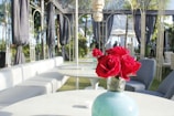 A beautifully arranged outdoor seating area features elegant round tables with plush seating. In the foreground, a vase filled with vibrant red roses adds a burst of color against the neutral tones of the furniture. The backdrop consists of an ornate metal structure draped with black curtains and surrounded by greenery, creating a sophisticated and inviting ambiance.