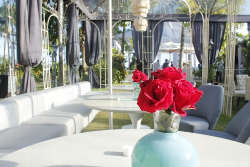 A beautifully arranged outdoor seating area features elegant round tables with plush seating. In the foreground, a vase filled with vibrant red roses adds a burst of color against the neutral tones of the furniture. The backdrop consists of an ornate metal structure draped with black curtains and surrounded by greenery, creating a sophisticated and inviting ambiance.