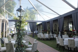 An outdoor venue with a glass roof featuring elegant dining arrangements. Round and rectangular tables are covered with white tablecloths, surrounded by padded chairs. A lamppost wrapped in greenery stands in the foreground. Gray drapes hang from the ceiling along the perimeter, framing lush greenery visible on the outside. Various potted plants and red floral arrangements add a touch of color.