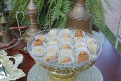 A decorative glass platter holds an assortment of Middle Eastern pastries, each resting in a small paper cup. The platter is set on an ornate golden stand. In the background, there are brass coffee pots and lush green ferns.