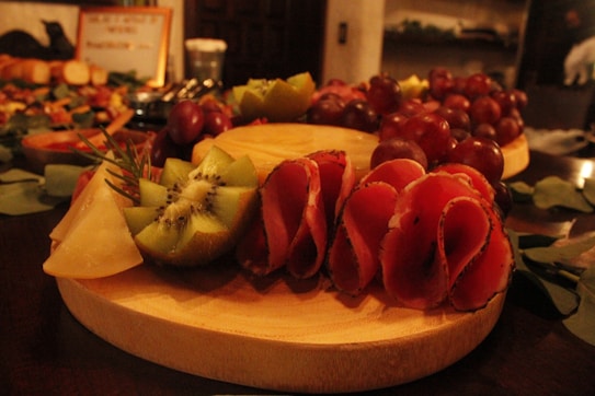 A wooden platter is beautifully arranged with a variety of foods including slices of kiwi, cheese wedges, and rolled cured meats. The platter is garnished with sprigs of rosemary, while clusters of red grapes are piled on the side. The setting suggests a warm, inviting atmosphere, possibly at a buffet or dinner party.
