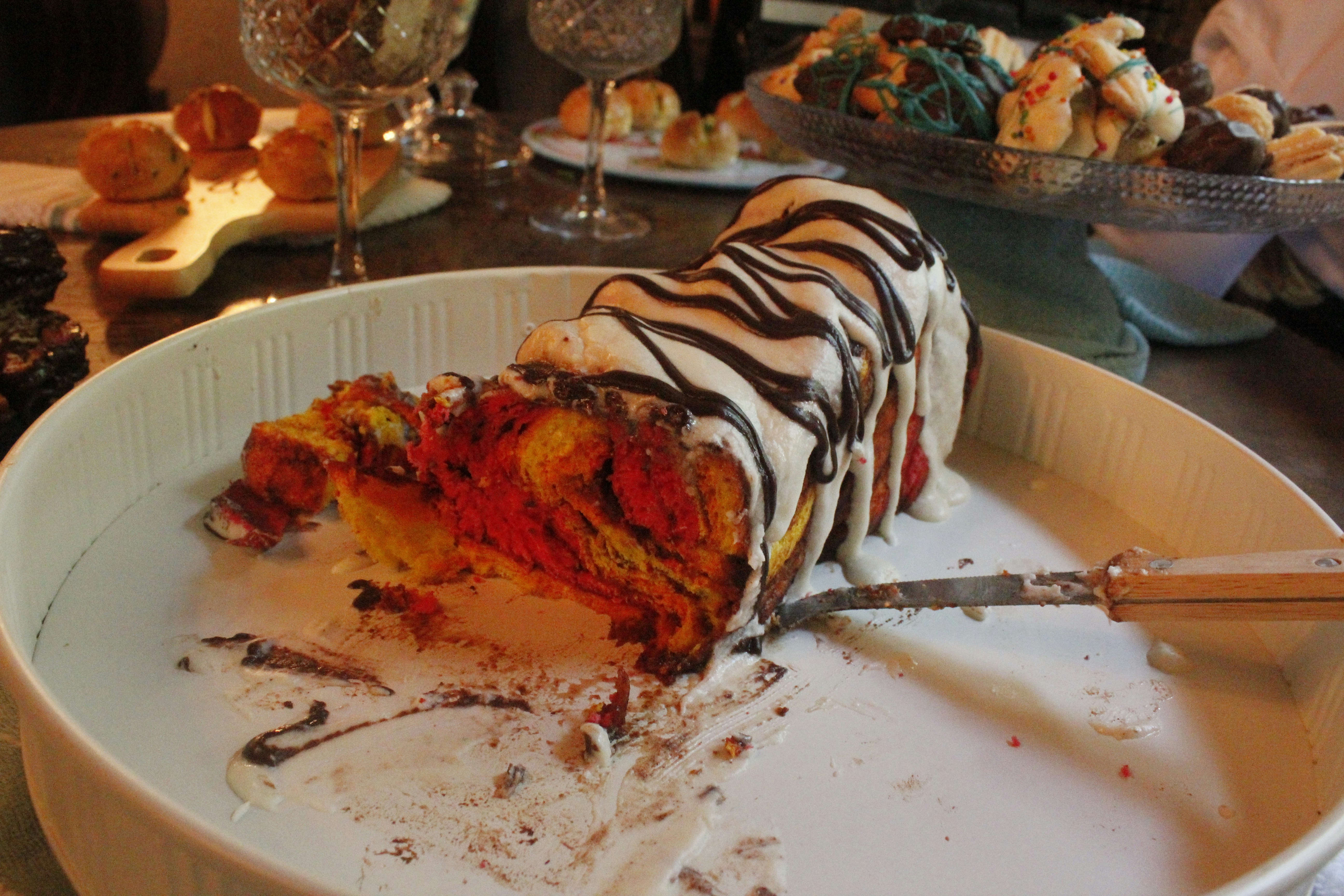 A richly colored dessert with swirls of red, yellow, and chocolate hues. The cake is topped with a drizzle of white and dark chocolate icing. It is placed in a large white bowl with a serving knife beside it. In the background, there are elegantly set wine glasses and a tray of various pastries and bread.