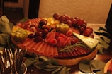 A charcuterie board filled with an assortment of meats, cheeses, grapes, walnuts, kiwi, and garnished with rosemary. The board is arranged artistically with slices of prosciutto and salami fanned out beside cubes of cheese and clusters of red grapes. A wedge of blue cheese is positioned prominently on the board.