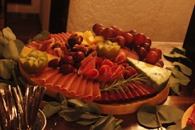 A charcuterie board filled with an assortment of meats, cheeses, grapes, walnuts, kiwi, and garnished with rosemary. The board is arranged artistically with slices of prosciutto and salami fanned out beside cubes of cheese and clusters of red grapes. A wedge of blue cheese is positioned prominently on the board.