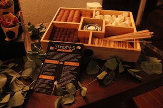 A wooden box contains separate compartments filled with marshmallows, graham crackers, chocolate, and a small can of fuel. Skewers are placed on top and a detailed guide for making s'mores is visible in front of the box. Green leaves are spread around the box for decoration.